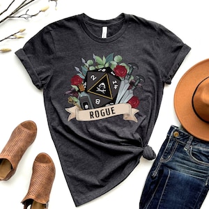 May include: A dark gray t-shirt with a graphic of a twenty-sided die with a skull and crossbones, surrounded by flowers, swords, and a potion bottle. The text "ROGUE" is on a banner below the die.