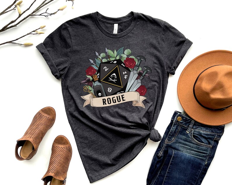 May include: A dark gray t-shirt with a graphic of a twenty-sided die with a skull and crossbones, surrounded by flowers, swords, and a potion bottle. The text "ROGUE" is on a banner below the die.
