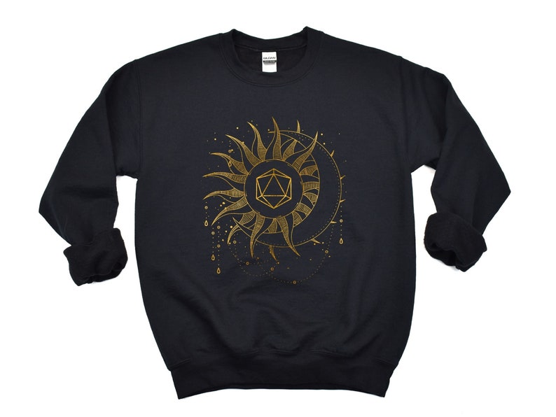 Puede incluir: Sudadera negra con un dise&ntilde;o dorado de sol y luna. El sol tiene rayos puntiagudos y una forma geom&eacute;trica en el centro. La luna tiene forma de media luna. L&iacute;neas punteadas y peque&ntilde;os c&iacute;rculos rodean el sol y la luna.