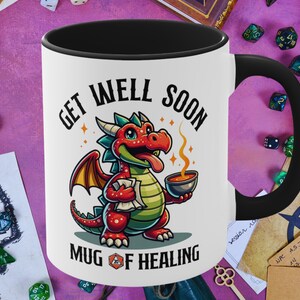 May include: White ceramic mug with a black handle. The mug has a cartoon illustration of a red dragon holding a steaming mug. The text "Get Well Soon Mug of Healing" is printed on the mug.