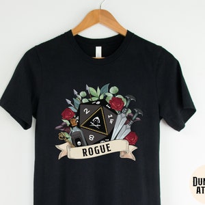 May include: Black t-shirt with a graphic of a 20-sided die, a vial, and daggers surrounded by flowers and leaves. The text "Rogue" is below the die.