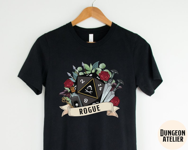 May include: Black t-shirt with a graphic of a 20-sided die, a vial, and daggers surrounded by flowers and leaves. The text "Rogue" is below the die.