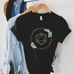May include: Black t-shirt with a gold geometric design and sprigs of green leaves. The design features a gold polygon inside a gold geometric frame. The t-shirt is hanging on a wooden hanger with other clothing.