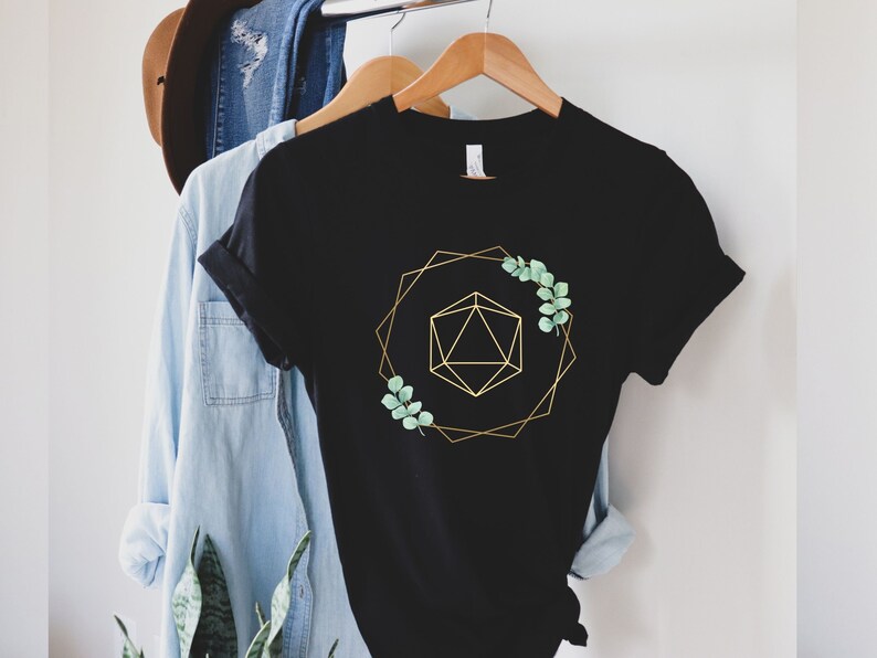 May include: Black t-shirt with a gold geometric design and sprigs of green leaves. The design features a gold polygon inside a gold geometric frame. The t-shirt is hanging on a wooden hanger with other clothing.