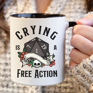 May include: White ceramic mug with a black rim and handle. The mug has a black and white illustration of a twenty-sided die with the text "Crying is a Free Action".