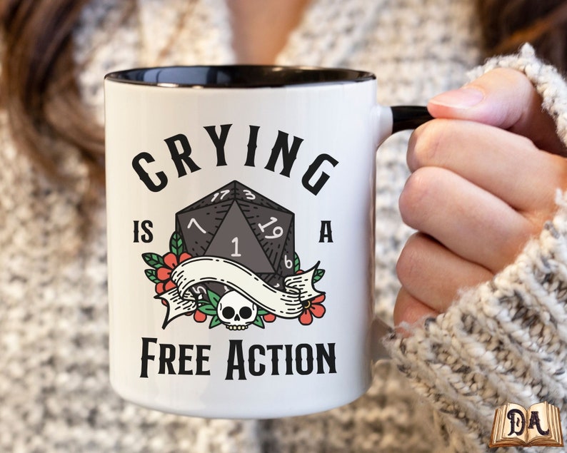 May include: White ceramic mug with a black rim and handle. The mug has a black and white illustration of a twenty-sided die with the text "Crying is a Free Action".