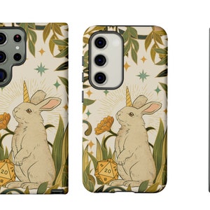May include: A phone case featuring a whimsical illustration of a unicorn rabbit with a golden horn, standing in a field with a 20-sided die. The design includes yellow flowers, green foliage, and a cream-colored background with stars.