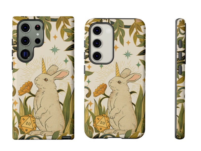 May include: A phone case featuring a whimsical illustration of a unicorn rabbit with a golden horn, standing in a field with a 20-sided die. The design includes yellow flowers, green foliage, and a cream-colored background with stars.