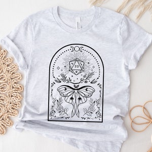 May include: A gray t-shirt with a black and white graphic design featuring a 20-sided die, a moth, and floral elements. The design is framed by a crescent moon and stars. The text "DUNGEON ATELIER" is visible in the bottom right corner.