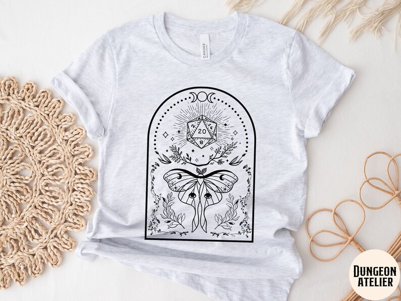 May include: A gray t-shirt with a black and white graphic design featuring a 20-sided die, a moth, and floral elements. The design is framed by a crescent moon and stars. The text "DUNGEON ATELIER" is visible in the bottom right corner.
