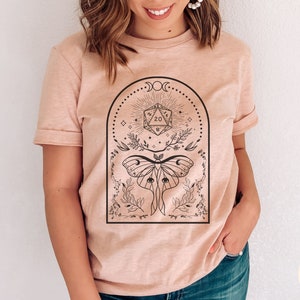 May include: A light pink t-shirt with a black and white graphic design featuring a moth, a 20-sided die, and floral elements. The graphic is surrounded by a crescent moon and stars.