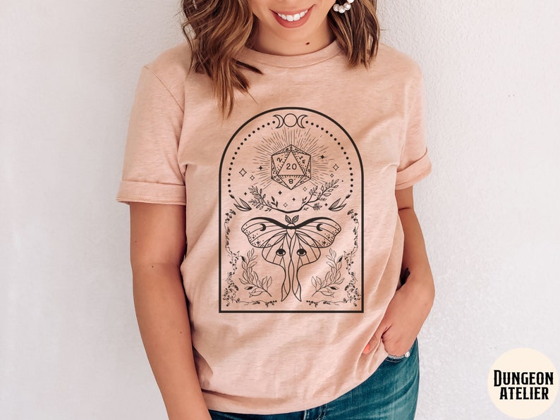 May include: A light pink t-shirt with a black and white graphic design featuring a moth, a 20-sided die, and floral elements. The graphic is surrounded by a crescent moon and stars.