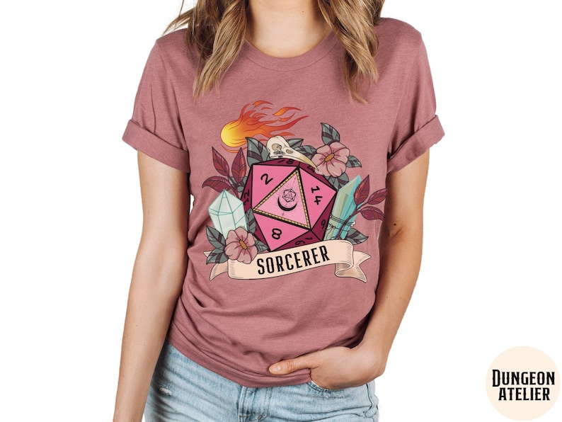 Puede incluir: Una camiseta rosa, roja y blanca con un gr&aacute;fico de un dado rosa de veinte caras con llamas, flores y una calavera. El texto "SORCERER" est&aacute; impreso en una pancarta debajo del dado.