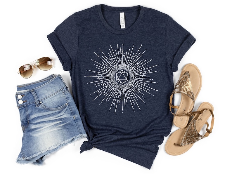 Puede incluir: Una camiseta azul marino con un gr&aacute;fico blanco de un dado de 20 caras rodeado de un dise&ntilde;o de rayos de sol. La camiseta se combina con pantalones cortos de mezclilla y sandalias doradas.