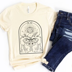 May include: A cream-colored t-shirt with a black graphic design featuring a moth, a twenty-sided die, and floral elements. The design is framed in a black arch with a moon and stars. The text "Dungeon Atelier" is visible in the bottom right corner.
