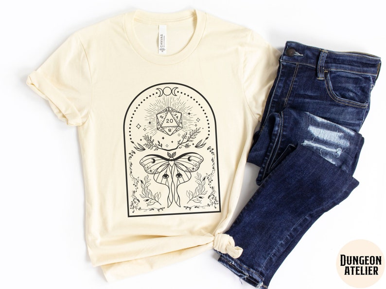 May include: A cream-colored t-shirt with a black graphic design featuring a moth, a twenty-sided die, and floral elements. The design is framed in a black arch with a moon and stars. The text "Dungeon Atelier" is visible in the bottom right corner.