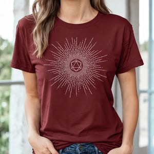 Puede incluir: Una camiseta color granate con un gr&aacute;fico blanco de un dise&ntilde;o de estallido de estrella geom&eacute;trico con un dado estilizado en el centro. El texto "Dungeon Atelier" es visible en la esquina inferior derecha de la imagen.