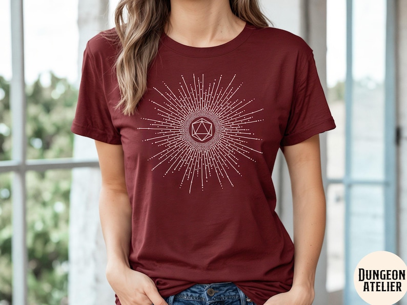 Puede incluir: Una camiseta color granate con un gr&aacute;fico blanco de un dise&ntilde;o de estallido de estrella geom&eacute;trico con un dado estilizado en el centro. El texto "Dungeon Atelier" es visible en la esquina inferior derecha de la imagen.