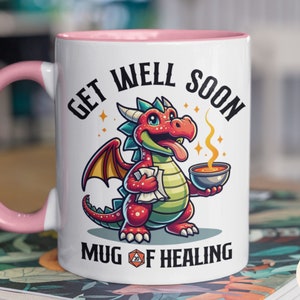 May include: White ceramic mug with a pink rim and handle. The mug features a cartoon red dragon holding a bowl of soup and a scroll. The text "Get Well Soon Mug of Healing" is printed on the mug.