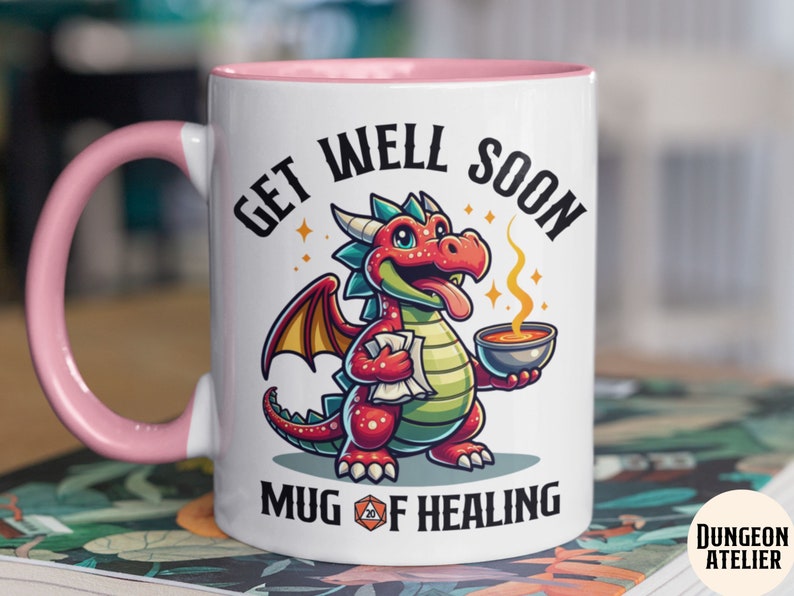 May include: White ceramic mug with a pink rim and handle. The mug features a cartoon red dragon holding a bowl of soup and a scroll. The text "Get Well Soon Mug of Healing" is printed on the mug.