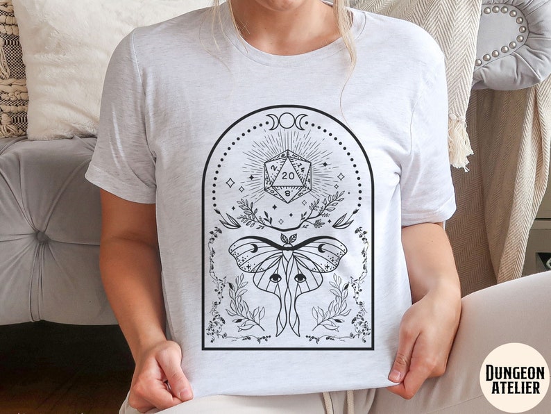 May include: White t-shirt with a black and white graphic design featuring a moth, a 20-sided die, and floral elements. The graphic is surrounded by a black circle with a crescent moon and stars.