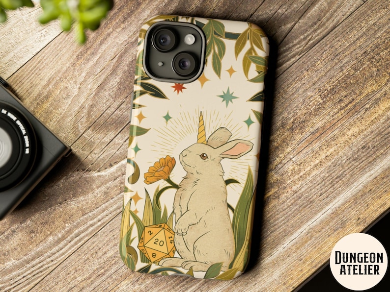 May include: A phone case featuring a whimsical illustration of a rabbit with a unicorn horn, surrounded by flowers, stars, and a D20 die. The case has a cream background with green and gold accents, and the text "Dungeon Atelier" is visible.