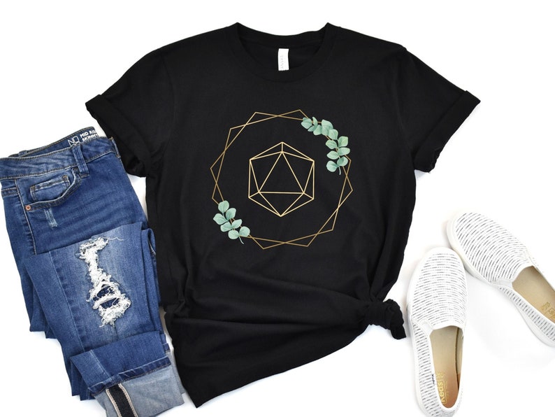 May include: Black t-shirt with a gold geometric design, a green leaf accent, and a geometric shape in the center. The shirt is paired with blue jeans and white slip-on shoes. The shirt is a casual style.