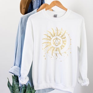 Puede incluir: Sudadera blanca con un dise&ntilde;o dorado de sol y luna. El gr&aacute;fico presenta una forma geom&eacute;trica en el centro del sol, con una luna creciente y l&iacute;neas punteadas. La sudadera est&aacute; colgada en una percha de madera.