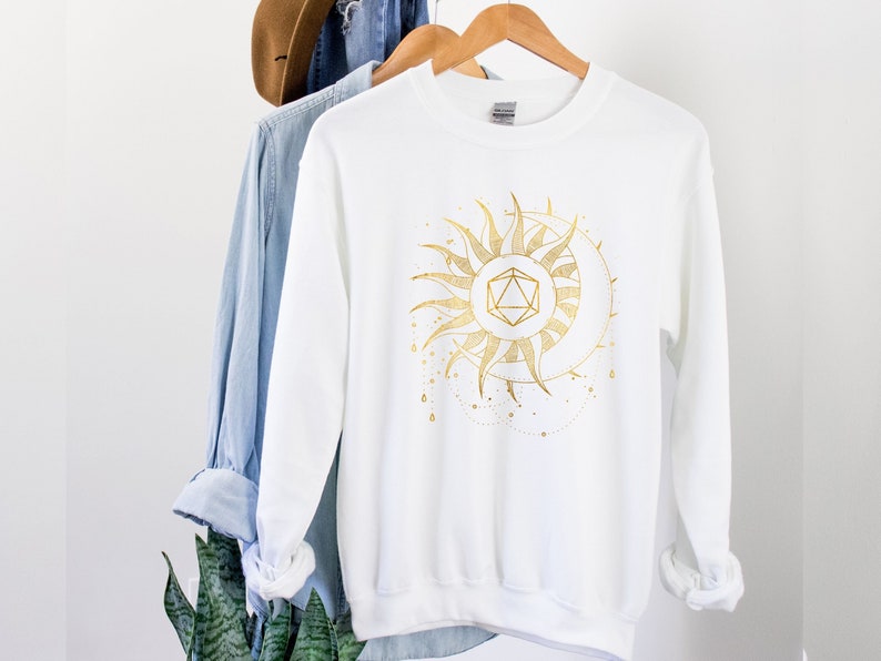 Puede incluir: Sudadera blanca con un dise&ntilde;o dorado de sol y luna. El gr&aacute;fico presenta una forma geom&eacute;trica en el centro del sol, con una luna creciente y l&iacute;neas punteadas. La sudadera est&aacute; colgada en una percha de madera.