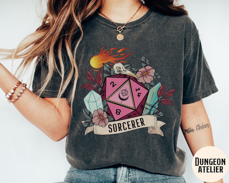 Puede incluir: Una camiseta gris oscuro con un gr&aacute;fico de dado de 20 caras rosa y blanco, rodeado de flores, cristales y llamas. El texto "SORCERER" est&aacute; impreso debajo del dado.