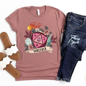 Puede incluir: Una camiseta rosa con un gr&aacute;fico de un dado de veinte caras rosa y rojo con llamas, una calavera y flores. El texto "SORCERER" est&aacute; debajo del dado.