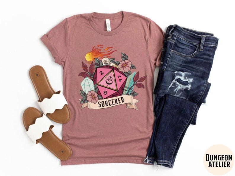 Puede incluir: Una camiseta rosa con un gr&aacute;fico de un dado de veinte caras rosa y rojo con llamas, una calavera y flores. El texto "SORCERER" est&aacute; debajo del dado.