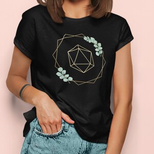 May include: Black t-shirt featuring a gold geometric design with a 20-sided die and sprigs of green leaves. The design is framed by a gold geometric shape. The model is wearing blue jeans.