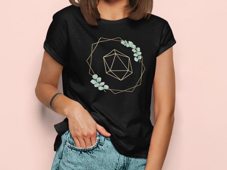 May include: Black t-shirt featuring a gold geometric design with a 20-sided die and sprigs of green leaves. The design is framed by a gold geometric shape. The model is wearing blue jeans.