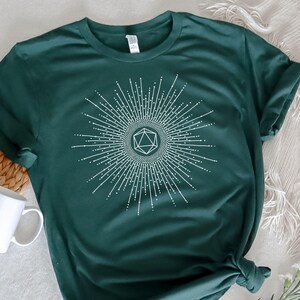 Puede incluir: Una camiseta verde oscuro con un gr&aacute;fico blanco de un dado de 20 caras dentro de un dise&ntilde;o de sol radiante. El texto "Dungeon Atelier" es visible en la esquina inferior derecha de la imagen.