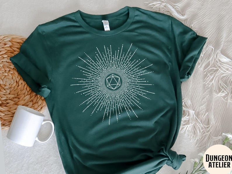 Puede incluir: Una camiseta verde oscuro con un gr&aacute;fico blanco de un dado de 20 caras dentro de un dise&ntilde;o de sol radiante. El texto "Dungeon Atelier" es visible en la esquina inferior derecha de la imagen.