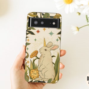 May include: A phone case featuring a whimsical illustration of a rabbit with a unicorn horn, surrounded by plants and a yellow 20-sided die. The design includes stars and a sunburst, with the text "Dungeon Atelier".