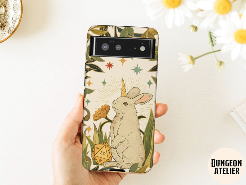 May include: A phone case featuring a whimsical illustration of a rabbit with a unicorn horn, surrounded by plants and a yellow 20-sided die. The design includes stars and a sunburst, with the text "Dungeon Atelier".