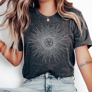 Puede incluir: Una camiseta gris oscuro con un gr&aacute;fico blanco de un dado de 20 caras rodeado de un dise&ntilde;o de rayos de sol. La camiseta est&aacute; hecha de un material suave y c&oacute;modo.
