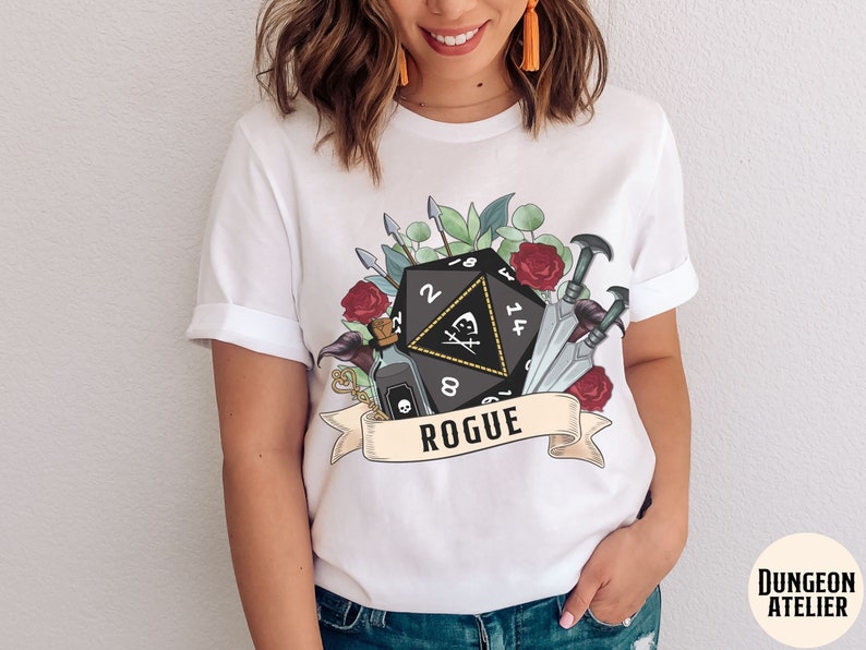 May include: White t-shirt with a black and white illustration of a 20-sided die, a vial, and daggers surrounded by flowers. The text "Rogue" is on a banner below the die.