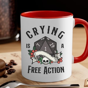 May include: White ceramic mug with a red rim and handle. The mug has a black and white illustration of a twenty-sided die with the text "Crying is a Free Action".