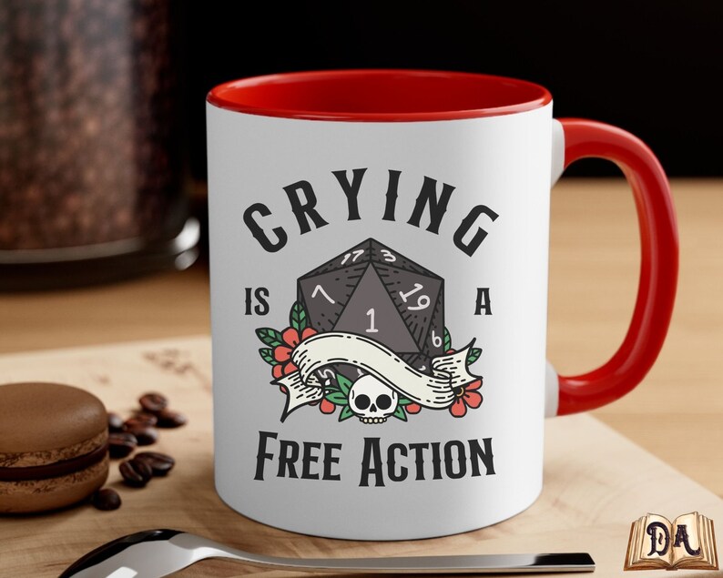 May include: White ceramic mug with a red rim and handle. The mug has a black and white illustration of a twenty-sided die with the text "Crying is a Free Action".