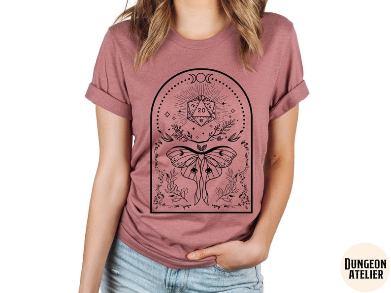 May include: A dusty rose t-shirt with a black graphic design featuring a moth, a twenty-sided die, and a crescent moon.
