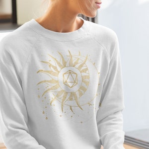 Puede incluir: Sudadera blanca con un dise&ntilde;o dorado de sol y luna con una forma geom&eacute;trica en el centro. El sol tiene rayos detallados y la luna tiene forma de media luna. El dise&ntilde;o tiene un tema celestial.