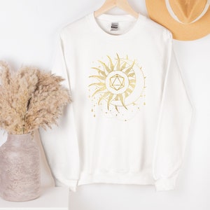 Puede incluir: Sudadera blanca con un dise&ntilde;o dorado de sol y luna. El gr&aacute;fico presenta una forma geom&eacute;trica en el centro, rodeada de rayos de sol y una luna creciente. El dise&ntilde;o tiene un tema celestial.