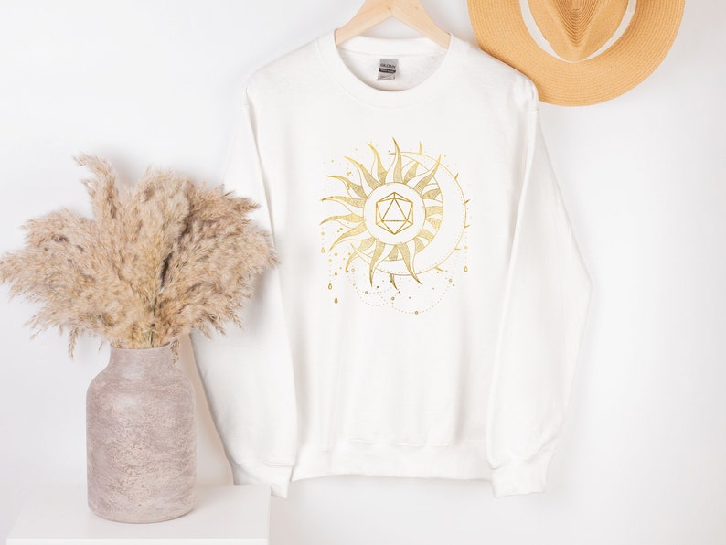 Puede incluir: Sudadera blanca con un dise&ntilde;o dorado de sol y luna. El gr&aacute;fico presenta una forma geom&eacute;trica en el centro, rodeada de rayos de sol y una luna creciente. El dise&ntilde;o tiene un tema celestial.