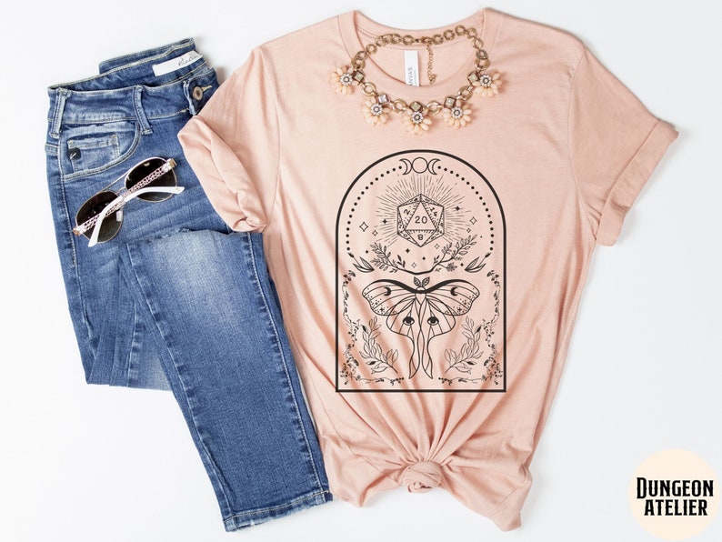 May include: A peach-colored t-shirt with a black and white graphic design featuring a moth, a twenty-sided die, and a crescent moon. The text "Dungeon Atelier" is visible in the bottom right corner of the image.