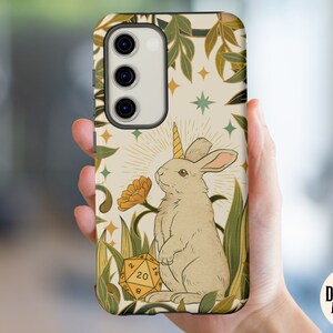 May include: A phone case featuring a whimsical illustration of a unicorn rabbit with a golden horn, standing near a yellow flower and a twenty-sided die. The design includes green foliage and stars on a cream background.