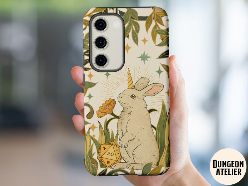 May include: A phone case featuring a whimsical illustration of a unicorn rabbit with a golden horn, standing near a yellow flower and a twenty-sided die. The design includes green foliage and stars on a cream background.