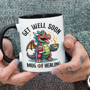 May include: White ceramic mug with a black handle. The mug has a cartoon illustration of a red and green dragon holding a bowl of soup. The text on the mug reads "Get Well Soon Mug of Healing".
